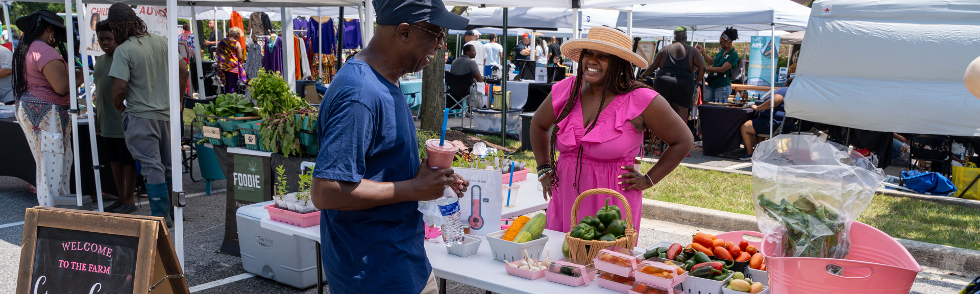 Community members visit vendors who display their vegetables and fruits on a table outdoors under a tent.