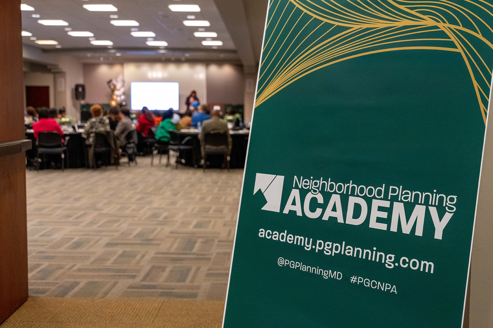 A sign in the foreground says, "Neighborhood Planning Academy, acandemy.pgplanning.com, @PGPlanningMD, #PGCNPA"; along with people in the background grouped at large round tables in front of a large screen.