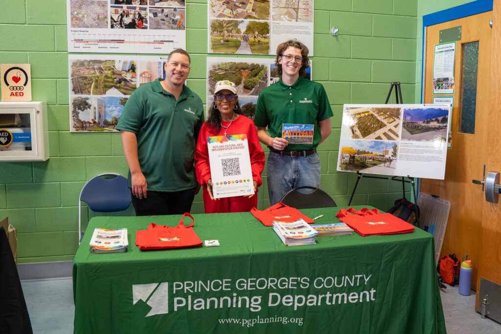 Prince George’s County Planning Department staffers stand behind an outreach table displaying brochures, tote bags, and project materials for the Suitland Cultural Arts Implementation Strategy. Project boards and planning concepts are displayed in the background.