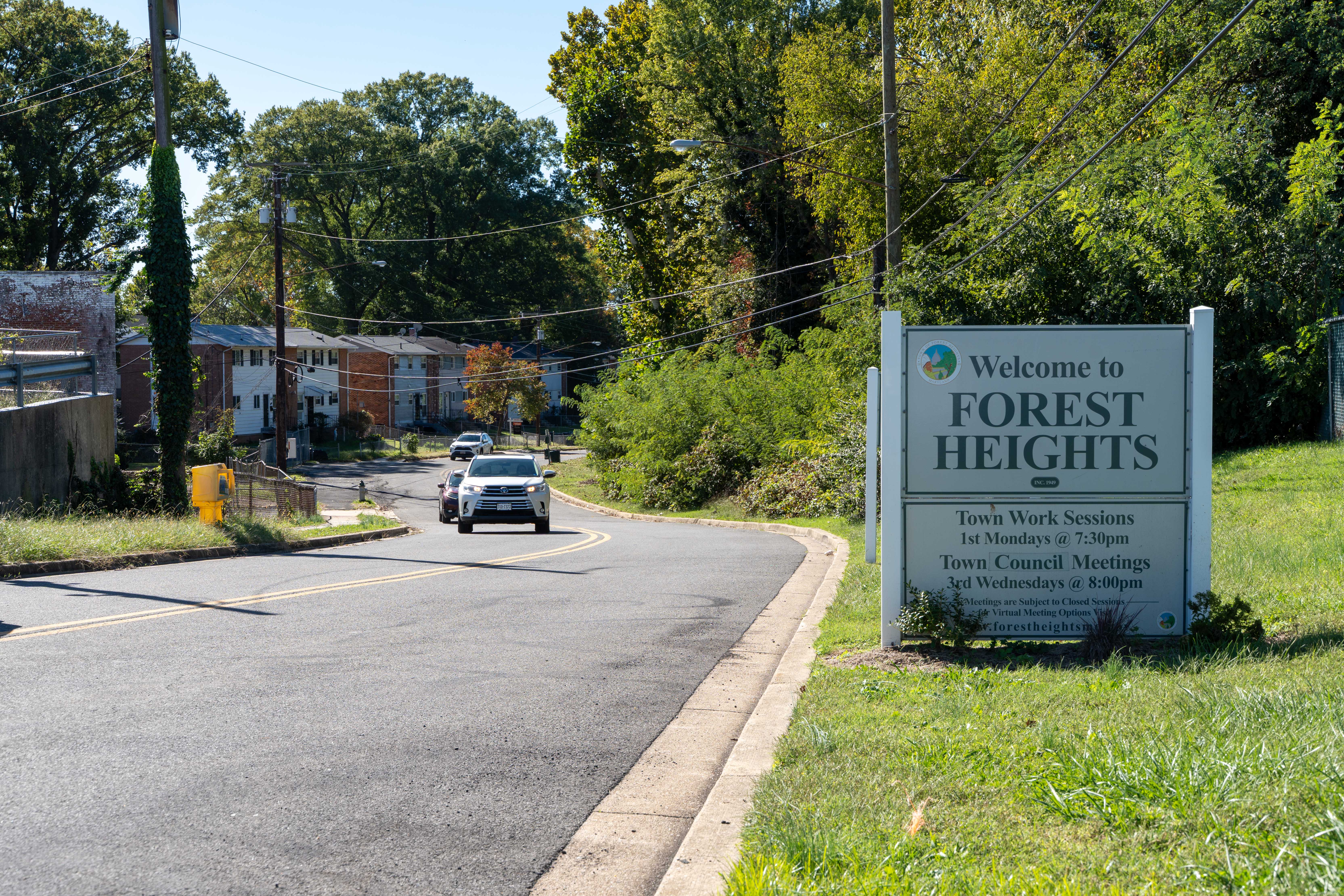 A sign for the town of Forest Heights alongside a road with a white car driving on it.